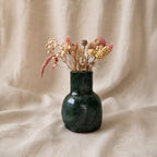 A small glossy dark green ceramic vase filled with dried flowers and grasses, set against a draped beige fabric background.