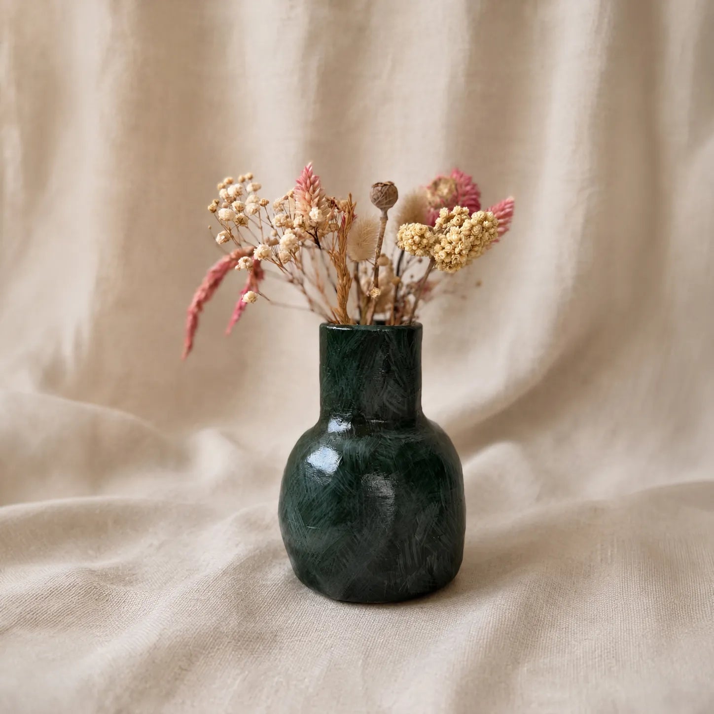 A small glossy dark green ceramic vase filled with dried flowers and grasses, set against a draped beige fabric background.