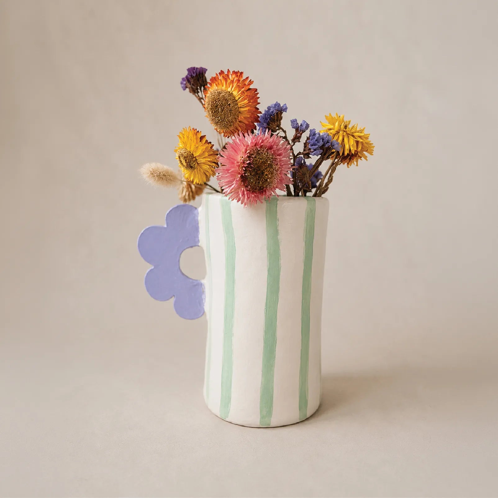 A white air dyr vase with vertical mint green stripes, holding a bouquet of dried multicolor flowers, set against a warm beige background.