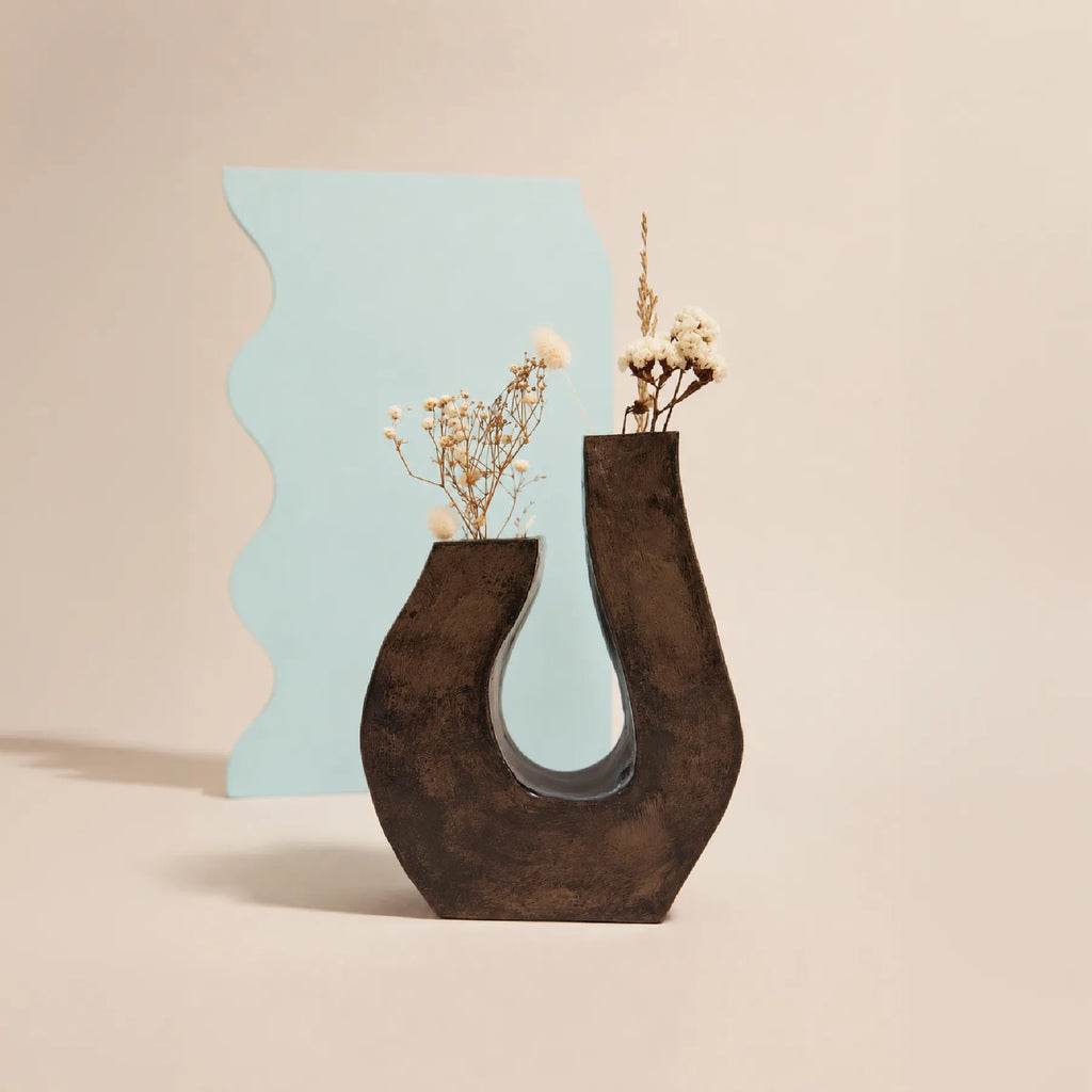 A sculptural dark brown pottery vase with a U-shaped cutout, styled with dried white flowers and a light blue wavy backdrop card on a warm beige background.