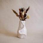 A textured off-white air dry vase with organic, flowing contours, filled with dried botanical stems and small flowers, set against a warm beige studio background.