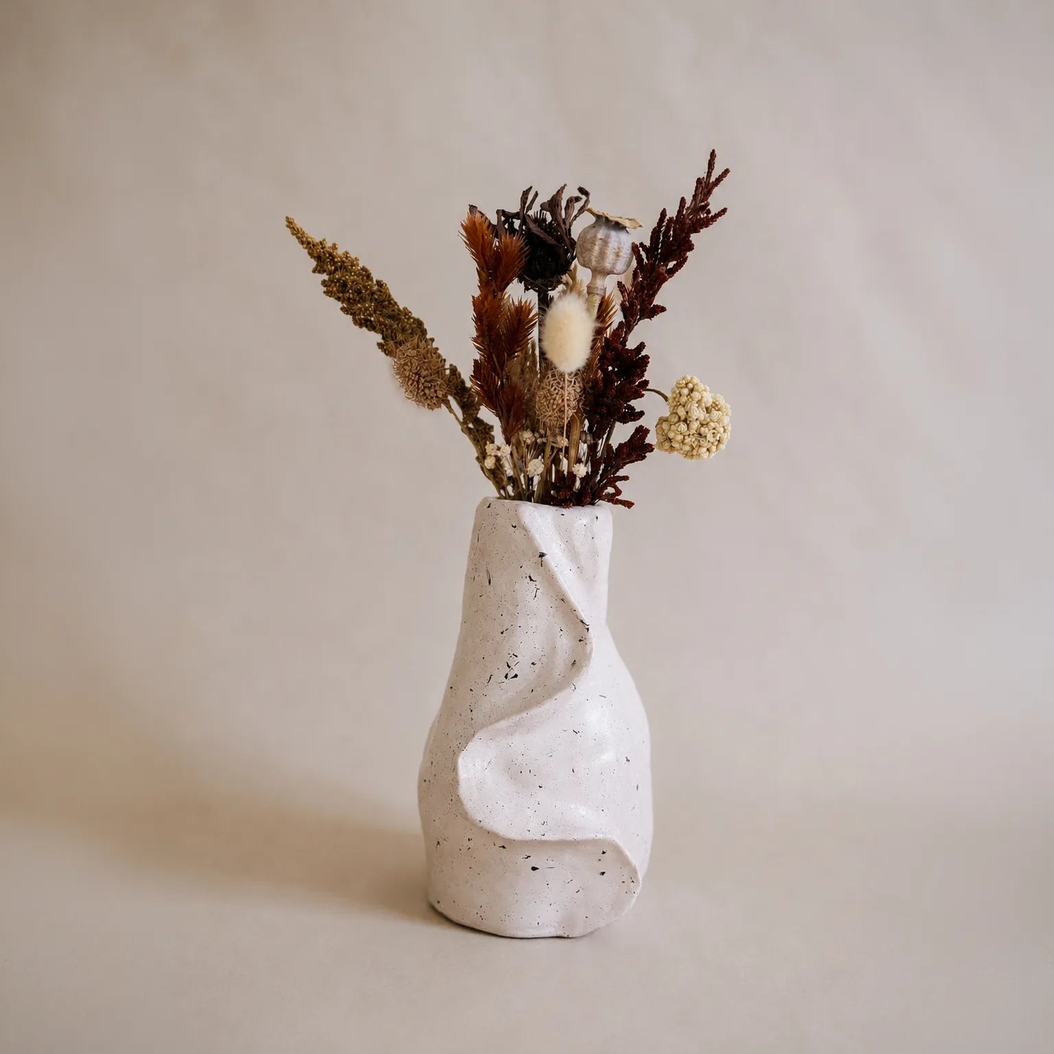 A textured off-white air dry vase with organic, flowing contours, filled with dried botanical stems and small flowers, set against a warm beige studio background.